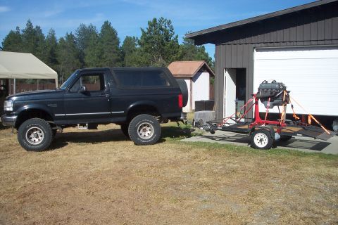1978 Ford Bronco - A black Ford Bronco is parked next to a garage, towing a trailer with an engine mounted on a stand.