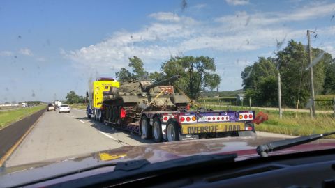 1996 Ford Bronco - A military tank is transported on a flatbed trailer down a highway, accompanied by other vehicles.