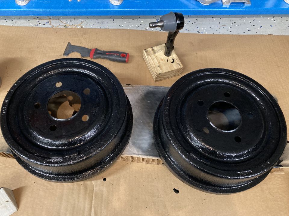 Two black brake drums sit on a cardboard-covered surface next to a scraper tool and an impact driver bit holder