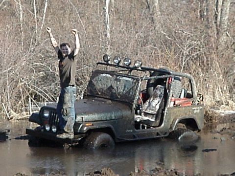 1992 Jeep Wrangler - A person celebrates while standing on a muddy Jeep Wrangler submerged in water, surrounded by trees.