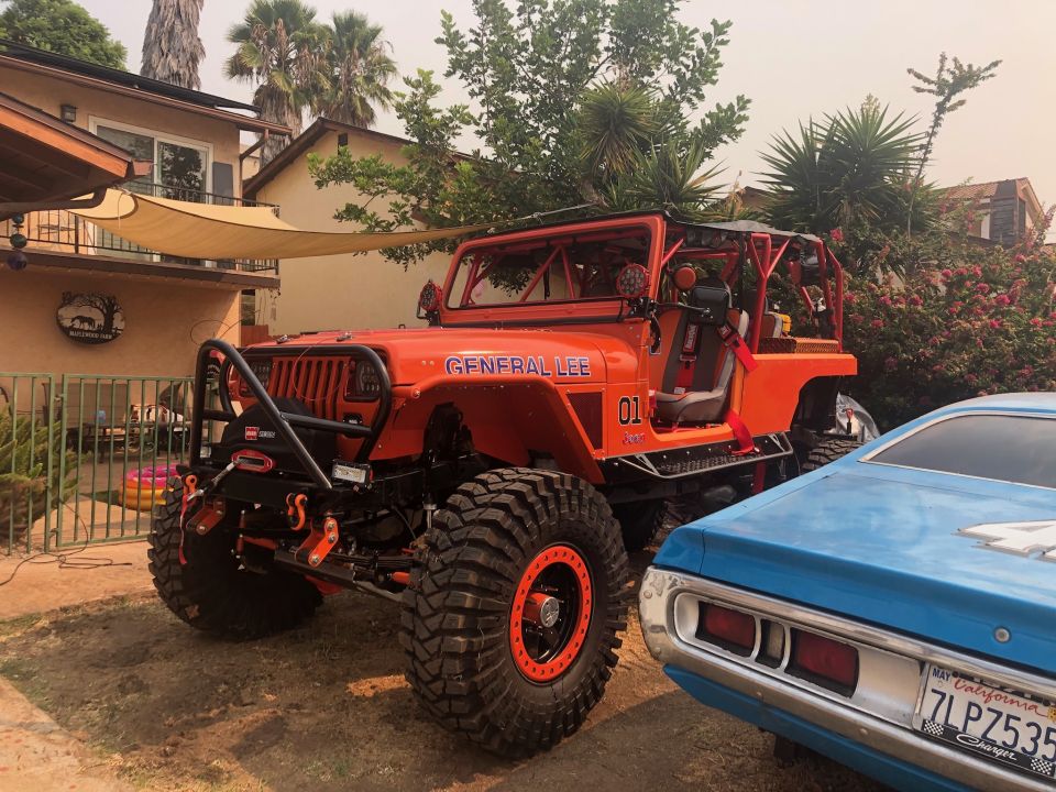 Orange Jeep Wrangler with 'GENERAL LEE' and '01' graphics, roll cage, large off-road tires, and winch. A blue car is parked next to it