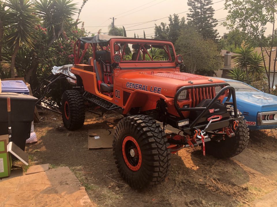 Orange Jeep Wrangler with "GENERAL LEE" and "01" on the hood, large off-road tires, and a winch on the front bumper. A blue car is parked next to it