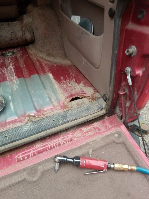 1994 Ford Bronco - The interior of a red vehicle shows significant rust damage on the floor, with an air ratchet nearby.