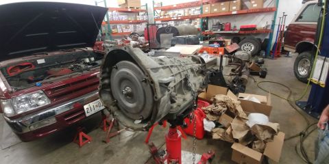 1996 Ford Bronco - A transmission is shown on a stand in a garage, with a red vehicle and other parts in the background.