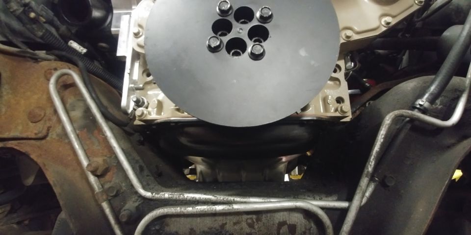 Close-up of a circular metal part with bolt holes, attached to an engine block with various hoses and metal lines visible