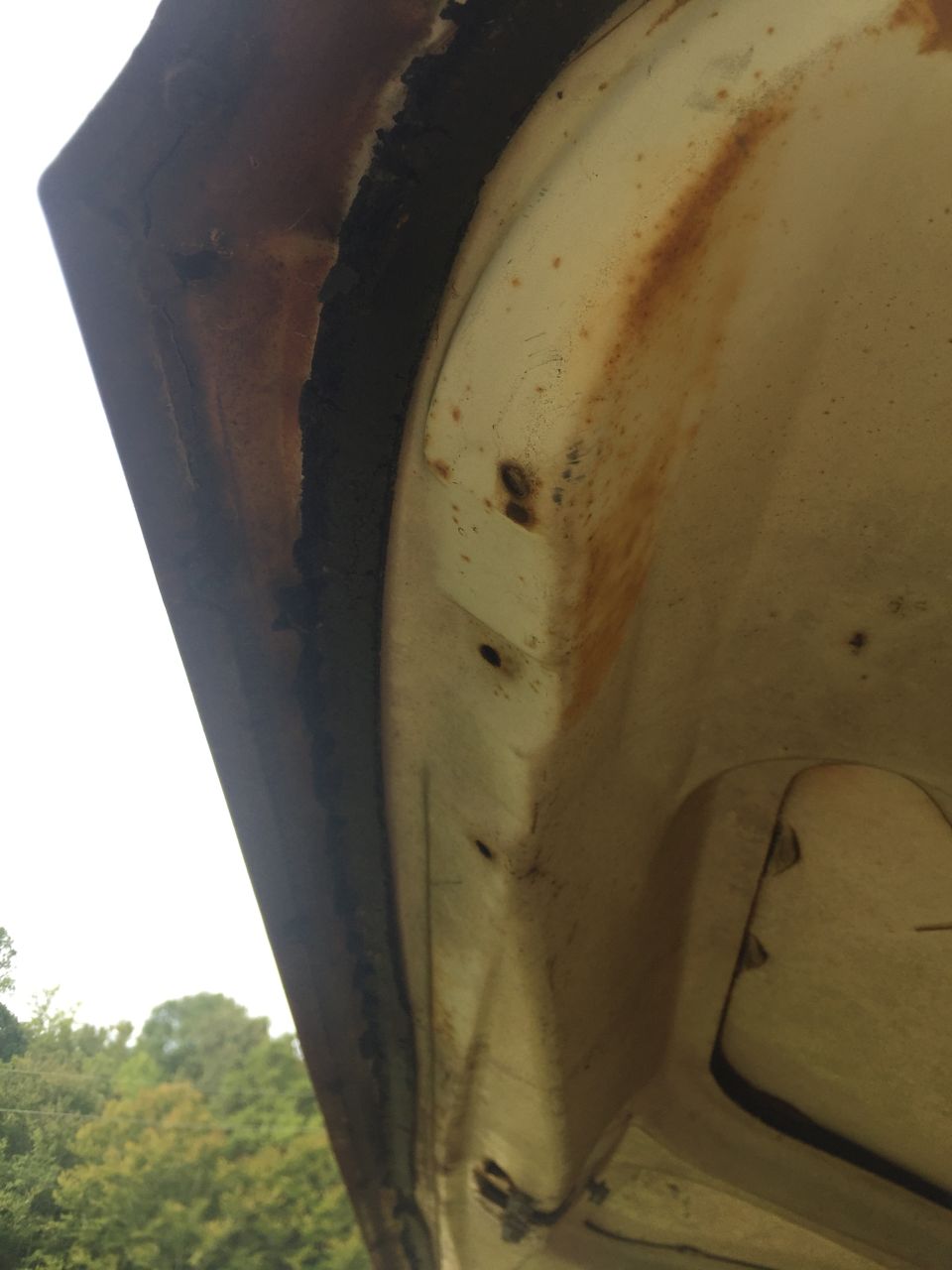 Underside of a cream-colored car hood showing rust and damage along the edge