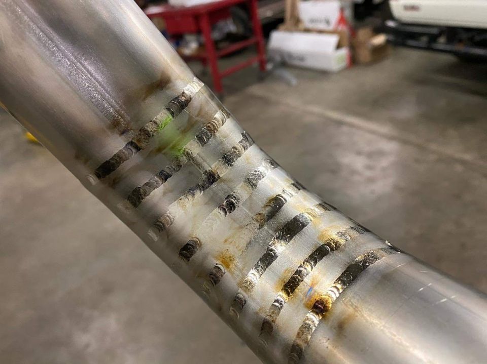 Close-up of a metal pipe with many welds, some with green residue. Supermotors.net logo visible