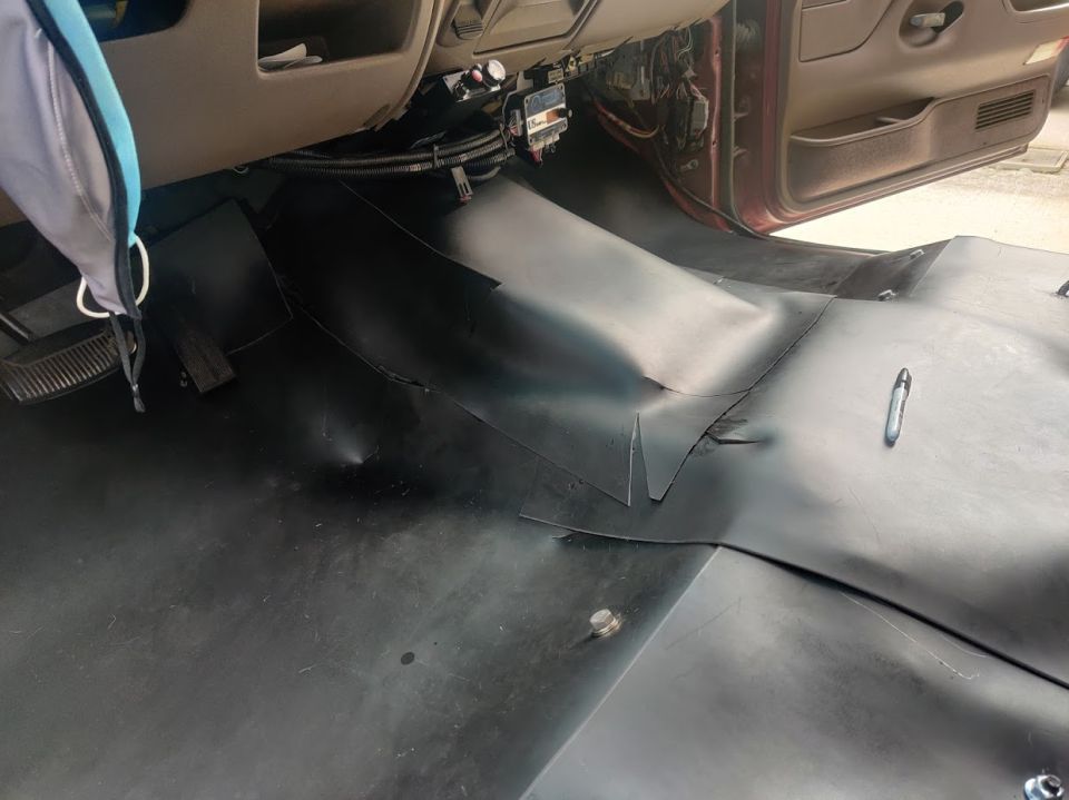 Interior floor of a vehicle with black rubber matting. Shows pedals, dashboard base, and door panel