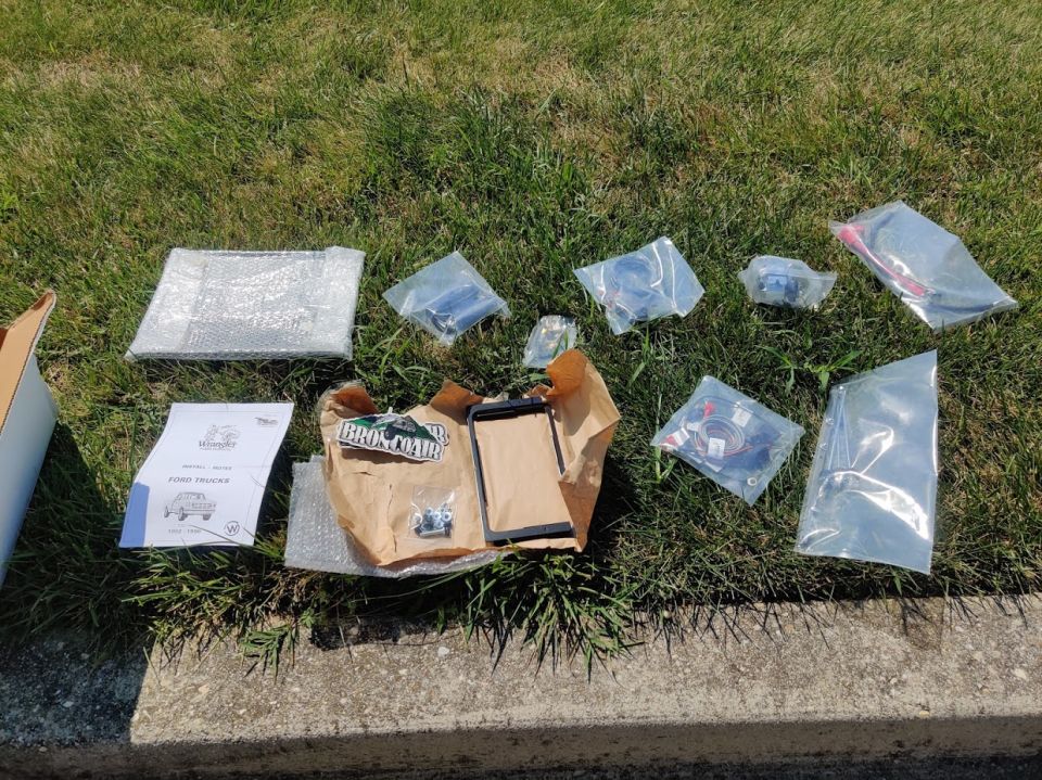 Various vehicle parts in plastic bags and bubble wrap, with a manual and a brown paper bag on grass