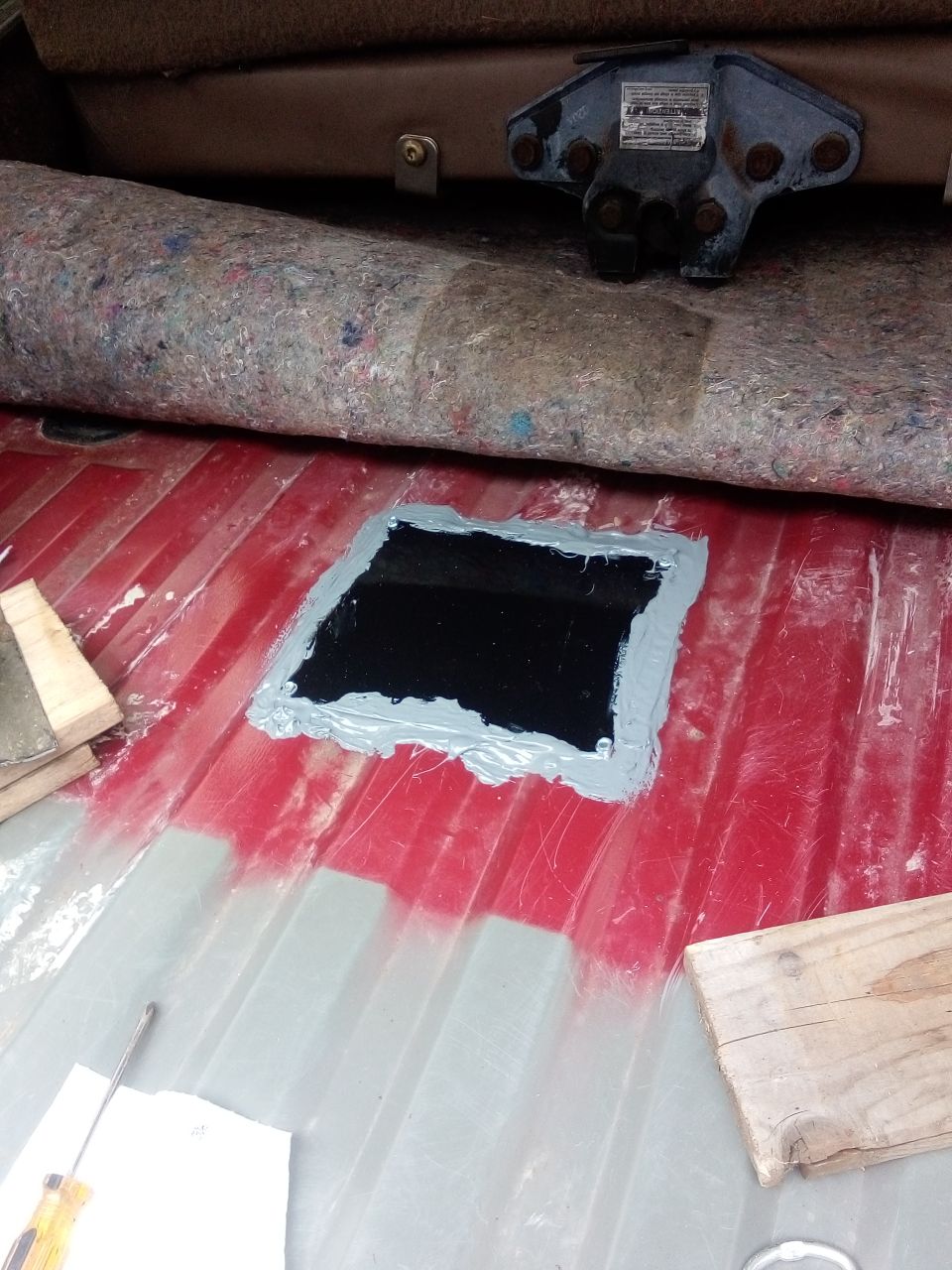 Patch being applied to the red floor of a vehicle's cargo area. A gray sealant surrounds a black square patch