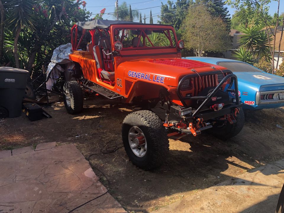 Orange Jeep Wrangler "General Lee" with "01" on the side, parked outdoors next to a blue car.