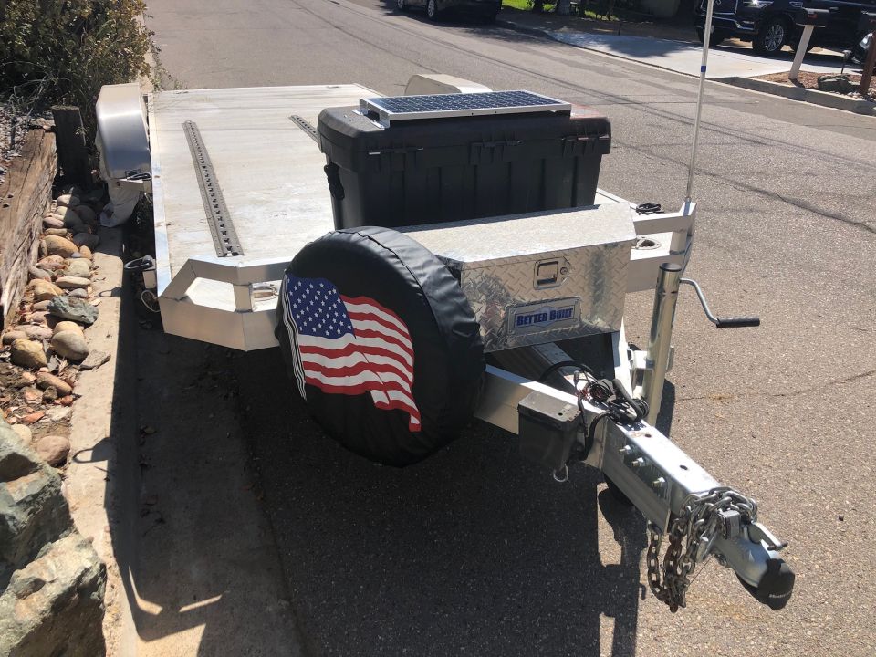 Utility trailer with American flag spare tire cover, toolbox, and solar panel.