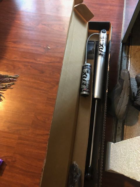 1992 Jeep Wrangler - Two silver Fox brand shock absorbers are shown inside a cardboard box, resting on a wooden floor. One shock absorber is longer than the oth…