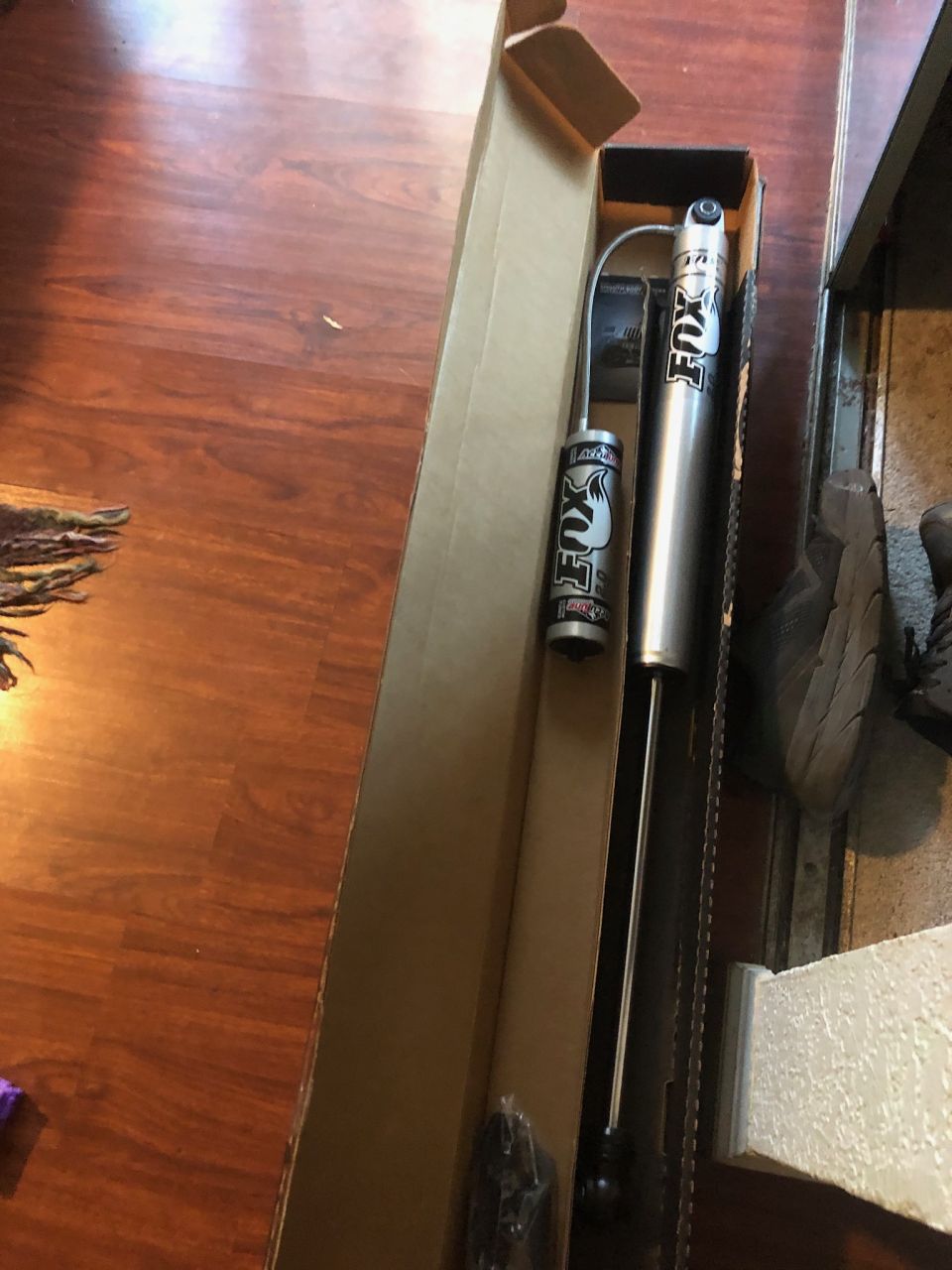 Two silver Fox shock absorbers in a box on a wooden floor.