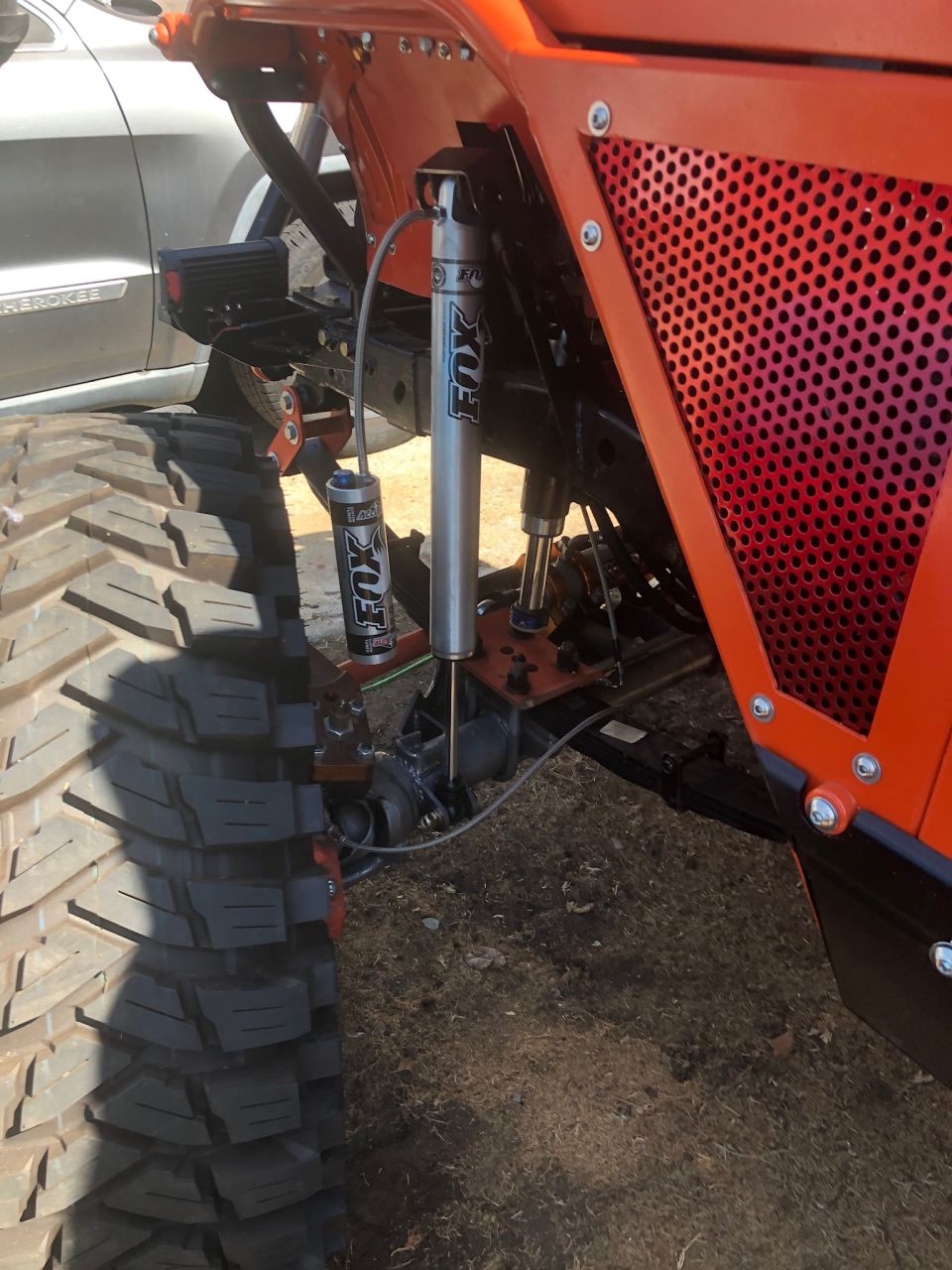 Modified Jeep Wrangler suspension with orange frame and Fox shocks next to a large off-road tire.