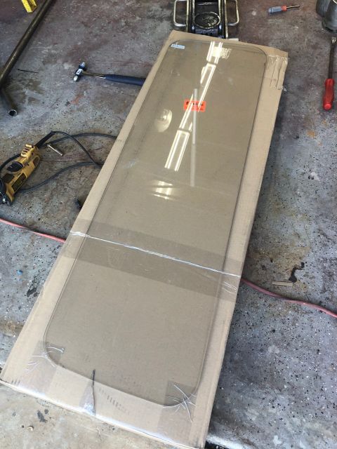 1992 Jeep Wrangler - A new, clear windshield is laid out on a cardboard box on a concrete floor. The windshield is wrapped in plastic and secured with tape.