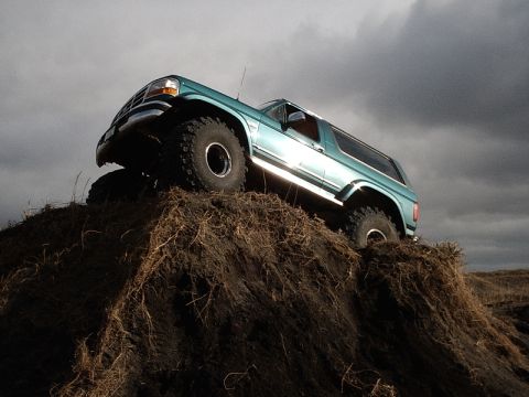 1996 Ford Bronco - A teal Ford Bronco navigates a steep dirt incline against a dramatic, overcast sky.