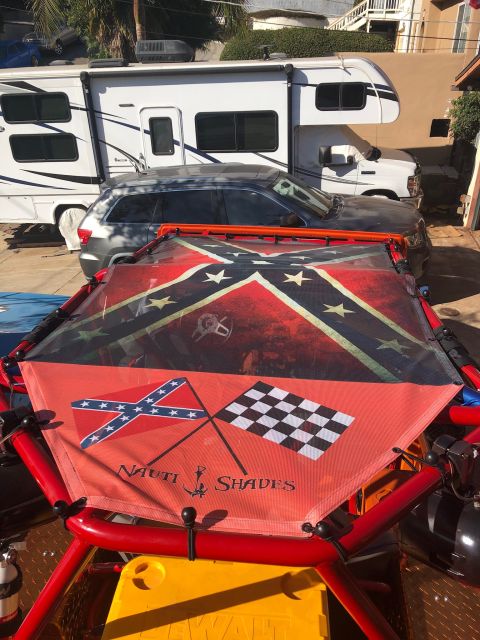 1992 Jeep Wrangler - A red metal frame surrounds a fabric canopy featuring a Confederate flag and a checkered flag, with the text "NAUTI SHADES" below. An RV an…