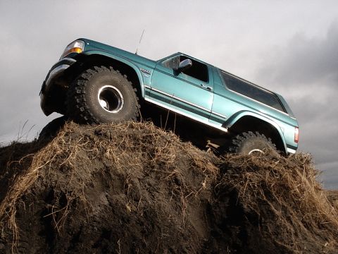 1996 Ford Bronco - A teal 1996 Ford Bronco navigates an off-road obstacle, its large tires gripping the loose dirt.