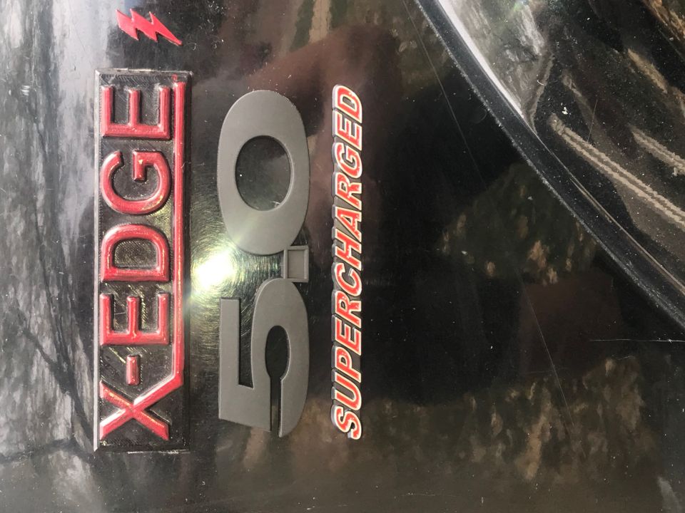 Close-up of X-EDGE 5.0 SUPERCHARGED text and red lightning bolt emblem on a black vehicle.