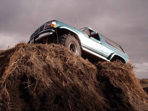 1996 Ford Bronco - A teal 1996 Ford Bronco is shown ascending a large dirt mound with sparse vegetation.