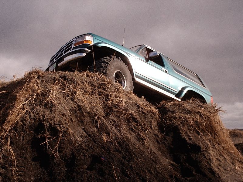 Teal Ford Bronco SUV on a dirt mound shot from low front-left angle under cloudy sky