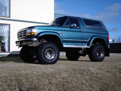 1996 Ford Bronco - A lifted teal 1996 Ford Bronco is parked on a grassy area next to a building.