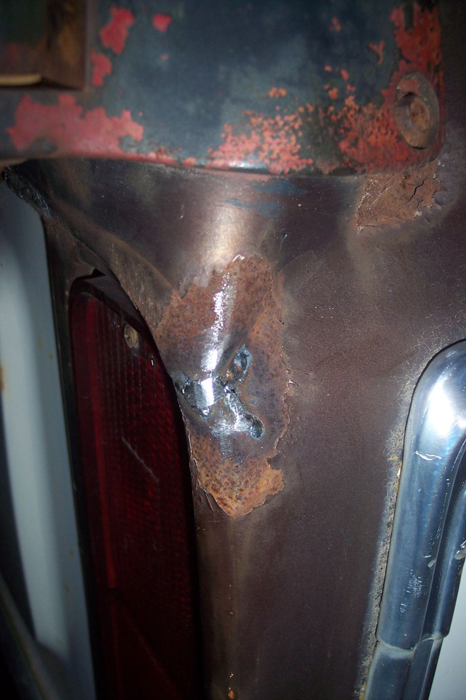 Close-up of rusted metal with welds near a red taillight on a vehicle.