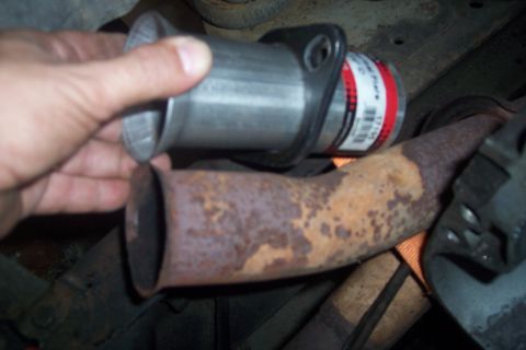 1978 Ford Bronco - A hand holds a new exhaust pipe adapter with a flange, preparing to connect it to a rusty, bent exhaust pipe section.