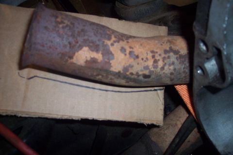 1978 Ford Bronco - A rusty exhaust pipe is shown resting on a piece of cardboard with a line drawn on it. A black strap is visible behind the pipe, and a dark…