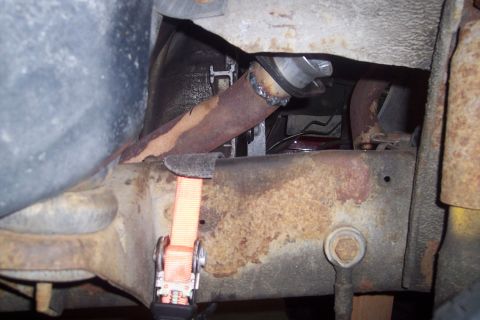 1978 Ford Bronco - A close-up view of the undercarriage of a vehicle, showing rusted exhaust pipes and frame components. An orange ratchet strap is used to se…