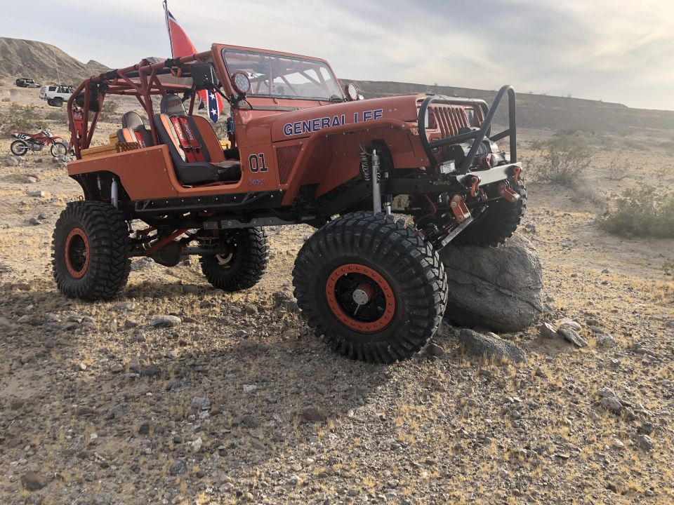 Orange Jeep Wrangler with roll cage, large tires, and "GENERAL LEE" text on fender, parked on rocky terrain