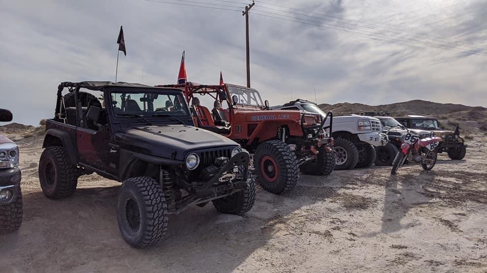 Black Jeep Wrangler with large tires parked on dirt, other off-road vehicles and a dirt bike behind it