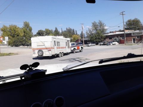 1978 Ford Bronco - A two-tone Ford Bronco tows a white travel trailer past buildings and trees under a clear blue sky.