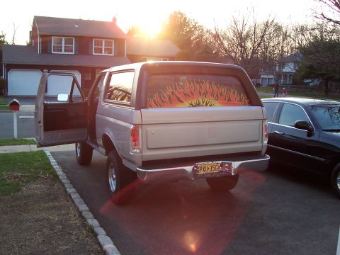 1990 Ford Bronco - A 1990 Ford Bronco is parked in a driveway with its door open, next to another vehicle. The rear window features a flame decal.