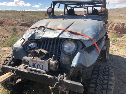 1992 Jeep Wrangler - A heavily damaged light blue Jeep Wrangler sits in a dry, outdoor landscape. The front end of the Jeep is severely crumpled, with an orange…