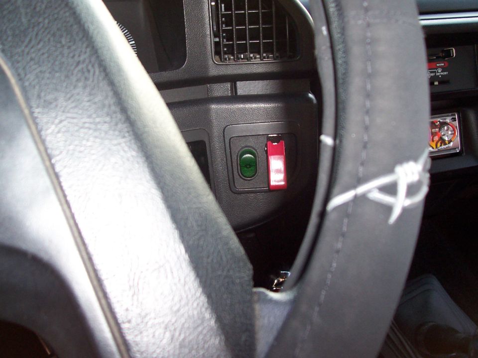Vehicle interior showing a green button and a red toggle switch on the dashboard, with part of the steering wheel visible