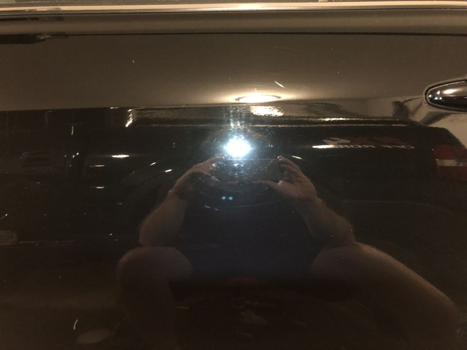 Reflection of a person holding a camera in the scratched black paint of a sedan.