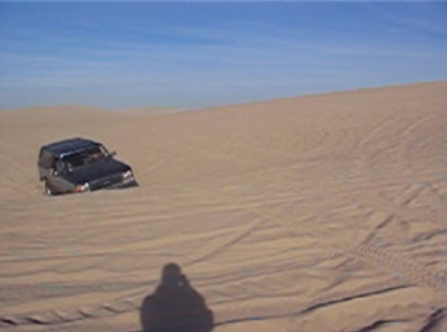 1989 Ford Bronco - A vehicle navigates a sandy terrain, its tires sinking into the soft ground under a clear blue sky.
