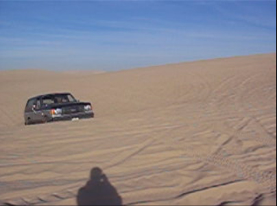 1989 Ford Bronco - A gray Ford Bronco navigates sandy terrain, kicking up dust under a bright blue sky.