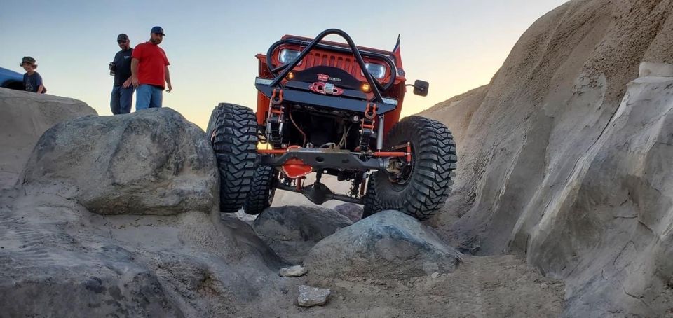 Orange Jeep Wrangler off-roading over rocks with people watching.