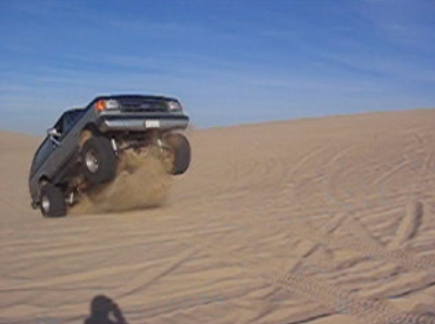 1989 Ford Bronco - A vehicle is captured mid-air as it jumps over a sand dune, with the tires kicking up a cloud of sand.