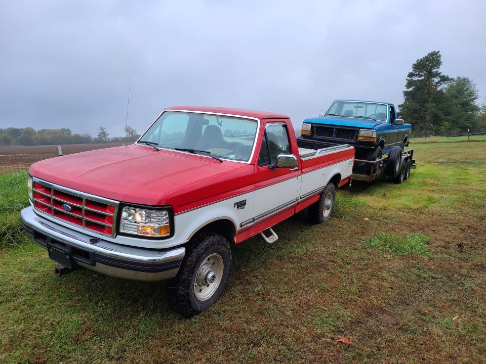 Red and white Ford pickup towing a blue Ford pickup on a trailer in a grassy field