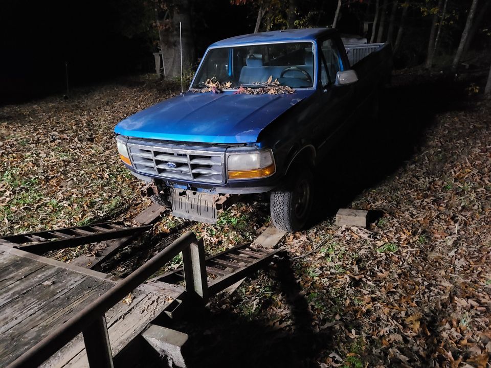 Blue Ford F-150 pickup at night with leaves on hood, missing front bumper, ramps in foreground