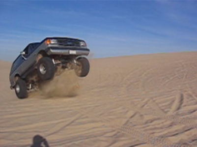 1989 Ford Bronco - SUV airborne over a sand dune, kicking up dust.