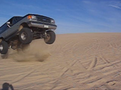 1989 Ford Bronco - A gray SUV soars through the air over sand dunes, with the "SUPERMOTORS" logo visible in the lower right corner.