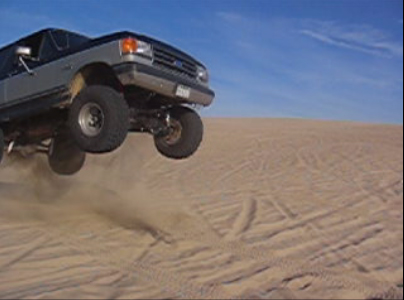 1989 Ford Bronco - Ford Bronco airborne over sand dunes, kicking up dust.