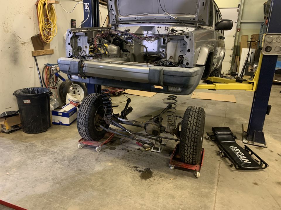 Silver Jeep Cherokee with front end disassembled, axle and suspension on dollies, in a garage.