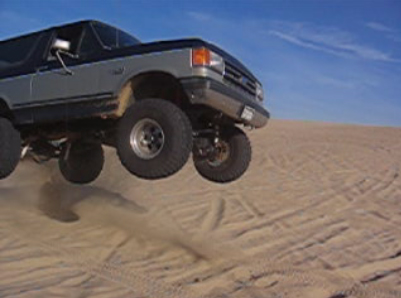1989 Ford Bronco - A lifted Ford Bronco soars through the air above sandy terrain under a clear blue sky.