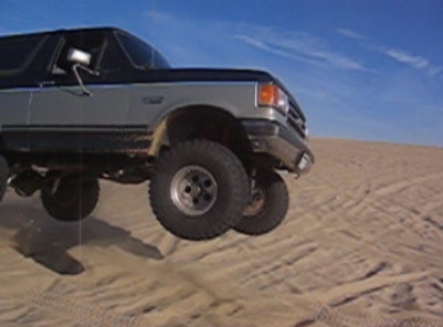 1989 Ford Bronco - A Ford Bronco jumps over sand dunes, captured mid-air against a clear blue sky.